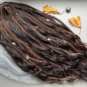 May include: Dark brown and light brown dreadlocks with decorative gold leaf and shell accents. The dreadlocks are styled with wavy ends and braided sections. The image includes fall foliage and a book in the background.