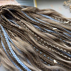 A close-up view of a collection of braided hair strands in various shades of brown and blue, with small white beads or pearls attached to some of the braids.