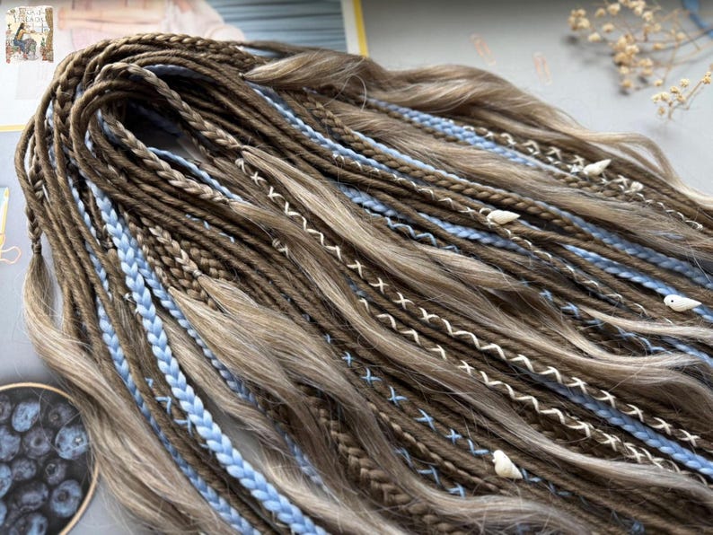 A close-up view of a collection of braided hair strands in various shades of brown and blue, with small white beads or pearls attached to some of the braids.