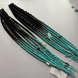 以下が含まれることがあります： 黒とターコイズのオームブレ合成ドレッドロックに、黒と白のアクセントが施されています。ドレッドロックはビーズとチャームで飾られています。