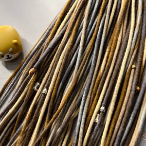 May include: Close-up of handmade dreadlocks in shades of brown, gray, and beige, adorned with wooden beads and metal charms. A yellow Shiba Inu figurine is visible in the upper left corner. The dreadlocks are arranged on a gray surface.