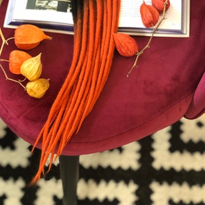 May include: A set of ombre red and black synthetic dreadlocks. The dreadlocks are lying on a burgundy velvet surface with a book and dried flowers.