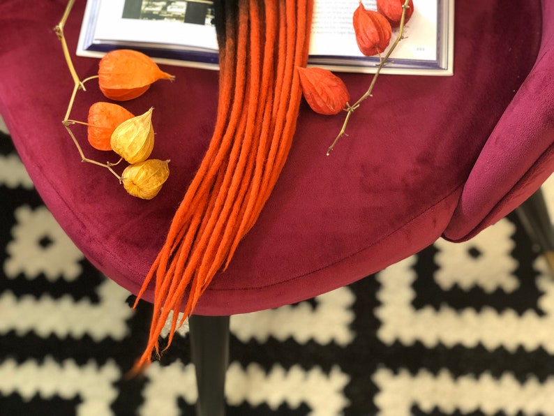 May include: A set of ombre red and black synthetic dreadlocks. The dreadlocks are lying on a burgundy velvet surface with a book and dried flowers.