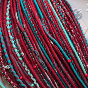 May include: A collection of red and teal dreadlocks. The dreads are a mix of solid colors and braided sections. Some dreads have small red beads and silver accents. The overall look is vibrant and textured.