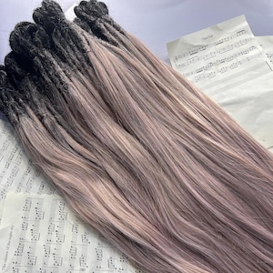 May include: A bundle of dreadlocks with a two-tone color scheme. The roots are dark, transitioning to a light pinkish-brown hue. The dreadlocks are lying on top of sheet music.