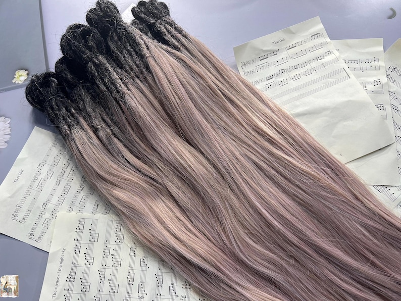 May include: A bundle of dreadlocks with a two-tone color scheme. The roots are dark, transitioning to a light pinkish-brown hue. The dreadlocks are lying on top of sheet music.