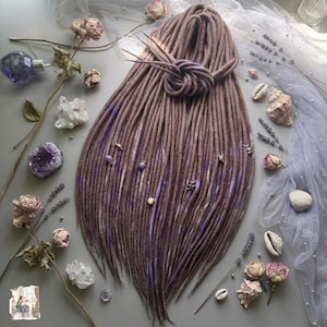 May include: A set of long, brown synthetic dreadlocks featuring subtle streaks of light purple and white. The dreadlocks are tied in a knot at the top and are adorned with small, metallic charms and beads in various shapes, including stars and spheres.