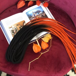 May include: A set of black and orange wool dreadlocks. The dreadlocks are arranged on a burgundy velvet surface with a book and dried flowers.