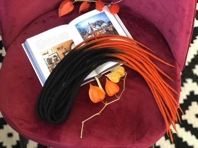 May include: A set of black and orange wool dreadlocks. The dreadlocks are arranged on a burgundy velvet surface with a book and dried flowers.