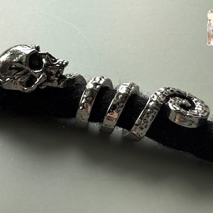 May include: Silver-colored dreadlock jewelry featuring a skull and spiral design. The skull is at the beginning of the spiral, which wraps around a black dreadlock. The jewelry has a hammered texture and is designed to be inserted into the hair.