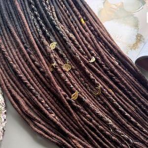 a collection of long, brown braided hair strands arranged on a surface, with some of the braids adorned with small gold or silver charms.