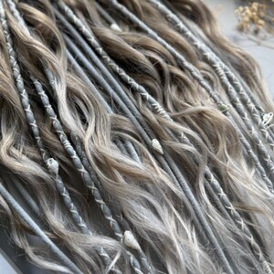May include: Close-up of a set of dreadlocks in shades of gray and light brown. The dreads are decorated with white stitching and small shell accents. The hair has a wavy texture and a natural appearance.