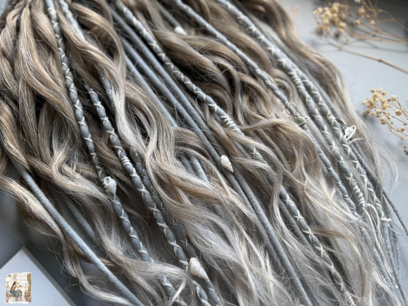 May include: Close-up of a set of dreadlocks in shades of gray and light brown. The dreads are decorated with white stitching and small shell accents. The hair has a wavy texture and a natural appearance.