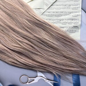 May include: A bundle of light brown hair extensions with subtle highlights. The hair is styled straight and lies across sheet music, a blue ribbon, and a pair of small scissors. The overall aesthetic suggests a focus on hair styling or beauty products.