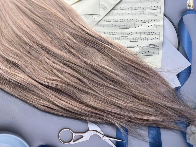 May include: A bundle of light brown hair extensions with subtle highlights. The hair is styled straight and lies across sheet music, a blue ribbon, and a pair of small scissors. The overall aesthetic suggests a focus on hair styling or beauty products.