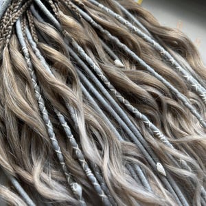 May include: Close-up of a set of dreadlocks in shades of brown and gray. The dreadlocks are styled with a mix of braids and wrapped sections, some with white stitching. The overall look is wavy and textured, with a natural appearance.