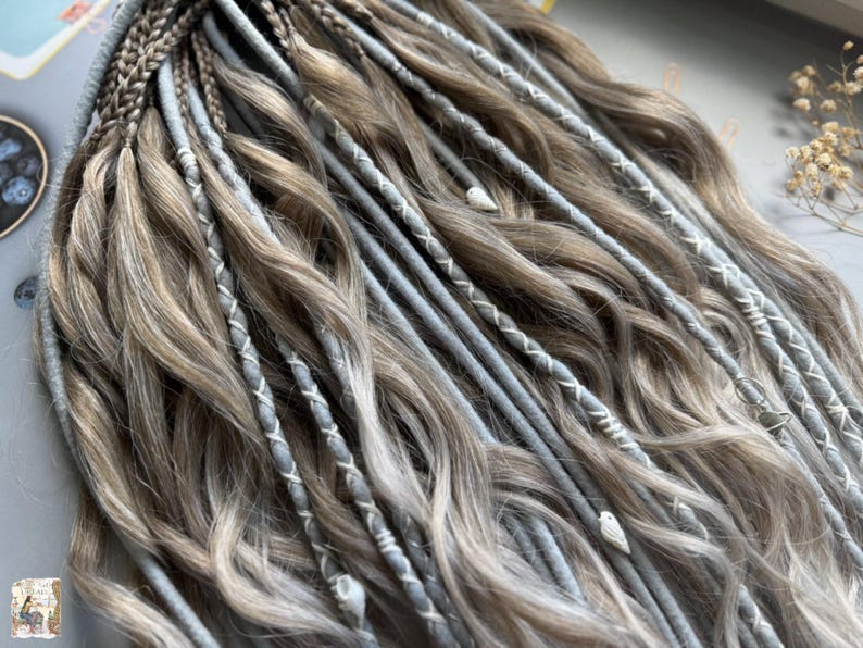 May include: Close-up of a set of dreadlocks in shades of brown and gray. The dreadlocks are styled with a mix of braids and wrapped sections, some with white stitching. The overall look is wavy and textured, with a natural appearance.