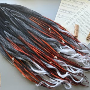 May include: A set of handmade dreadlocks in black, gray, and orange, with white wavy ends. The dreads are adorned with small silver charms. The image also includes sheet music and cinnamon sticks.