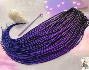 Könnte beinhalten: Ein Satz synthetischer Dreadlocks in Violett- und Schwarztönen. Die Dreadlocks liegen lose auf einem rosa und weißen Hintergrund.