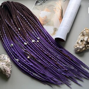 Peut inclure: Un ensemble de dreadlocks synthétiques présentant un dégradé de couleurs allant du brun foncé aux racines au violet vif aux pointes. Les dreadlocks sont ornées d'enroulements de fil argenté, de petites perles blanches et de minuscules coquillages.