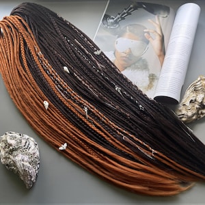 May include: A set of synthetic hair extensions featuring a gradient of dark brown to copper orange tones. The extensions consist of a mix of smooth dreadlocks and braided strands, accented with small silver-toned metal charms and white seashell beads.
