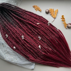 May include: Burgundy and black ombre dreadlocks with silver charms. The dreadlocks are decorated with mushroom, shell, and crescent moon charms. The hair is styled with a gradient effect, transitioning from black to burgundy.