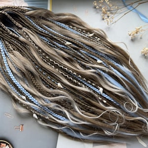 May include: Handmade dreadlocks in shades of brown, beige, and light blue. The dreads are styled with intricate braids, white stitching, and small shell accents. A unique hair accessory for a bohemian or alternative style.