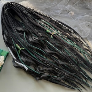 Puede incluir: Dreadlocks negros y grises con detalles verdes y blancos. Las rastas están adornadas con amuletos verdes en forma de pez. El cabello presenta una mezcla de texturas y colores, creando un aspecto único y artístico. Las rastas están dispuestas sobre una superficie gris claro.