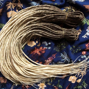 Pode incluir: Um pacote de dreadlocks sintéticas em dois tons de marrom, com a metade superior de uma cor loira mais clara. As dreadlocks estão dispostas em círculo sobre um tecido floral azul.