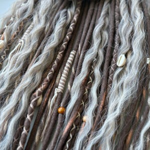 May include: Close-up of handmade dreadlocks in shades of brown and grey, with wavy texture. Some dreads are adorned with decorative stitching, beads, and seashells. The dreadlocks are a unique hair accessory.