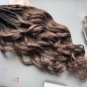 May include: A bundle of brown and black dreadlocks with wavy ends. The hair transitions from dark at the roots to a lighter brown. The dreadlocks are styled to appear natural and flowing. A silver perfume bottle is in the background.