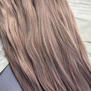 May include: Close-up of bundles of light brown hair extensions with subtle highlights. The hair is arranged on top of sheet music, creating a soft, textured appearance. The color is a blend of light brown and ash tones.