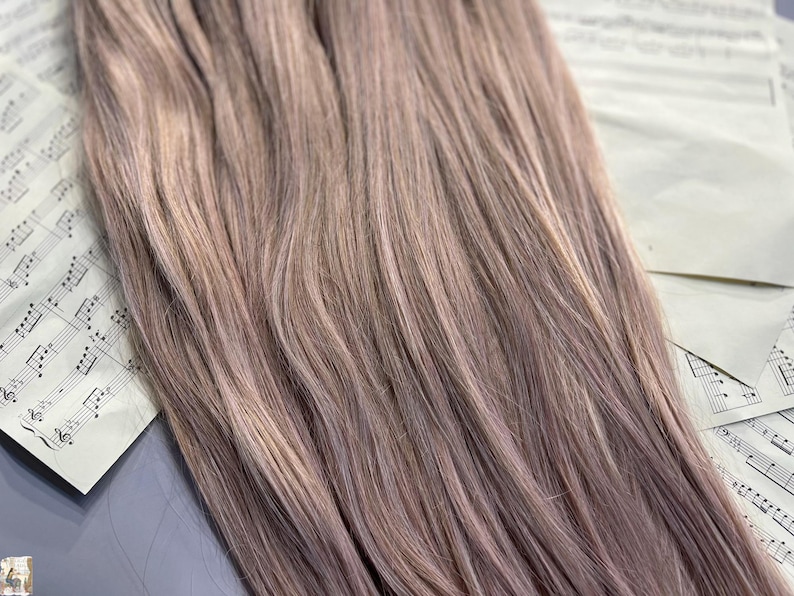 May include: Close-up of bundles of light brown hair extensions with subtle highlights. The hair is arranged on top of sheet music, creating a soft, textured appearance. The color is a blend of light brown and ash tones.