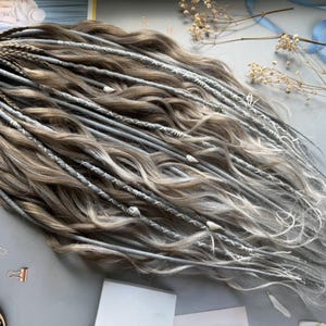 May include: A collection of handmade dreadlocks in shades of brown and gray, with some strands featuring white accents. The dreadlocks are styled with a wavy texture and adorned with decorative elements. A stylish hair accessory.