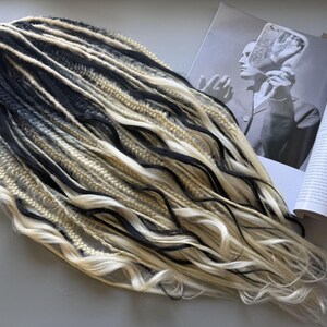 May include: A set of black and blonde synthetic dreadlocks. The dreads are arranged in a flowing pattern, with a mix of dark and light strands. A magazine and a small illustration are in the background.