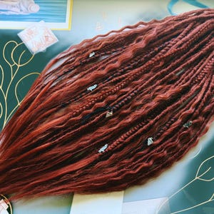 May include: A collection of reddish-brown dreadlocks, featuring diverse textures and braids. The dreads are decorated with small silver charms. The hair is arranged in a flowing style, with darker roots and lighter ends. The overall look is a bohemian, natural hairstyle.