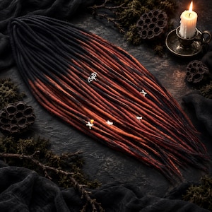 May include: A set of synthetic dreadlocks featuring a black to dark red ombre color gradient. The dreadlocks are adorned with small silver-toned charms, including a bat, bones, and other shapes, along with a single orange bead.