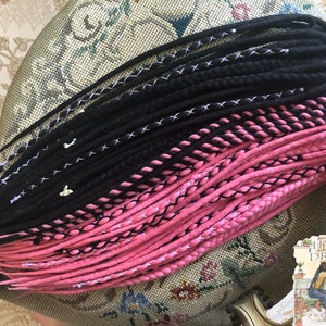 May include: A set of black and pink wool dreadlocks. The dreadlocks are arranged in two rows, with the black dreadlocks on top and the pink dreadlocks on the bottom. The dreadlocks are tied together with white string.
