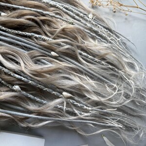 May include: Close-up of a collection of gray and light brown dreadlocks. The dreadlocks are adorned with white stitching and small decorative elements. The hair has a wavy texture and is arranged in a layered pattern.