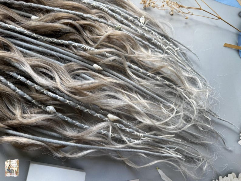 May include: Close-up of a collection of gray and light brown dreadlocks. The dreadlocks are adorned with white stitching and small decorative elements. The hair has a wavy texture and is arranged in a layered pattern.