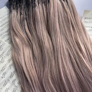 May include: A bundle of long, light brown hair extensions with dark roots. The hair is styled in straight strands and has a subtle ombre effect. Sheet music with the text "That Girl" is visible in the background.