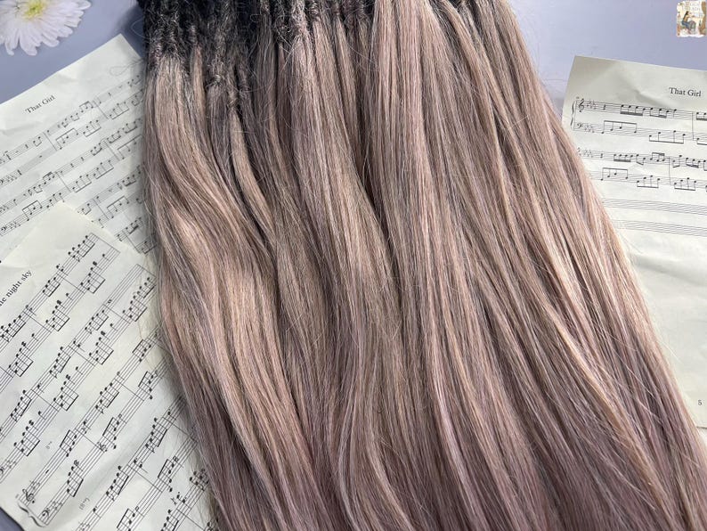 May include: A bundle of long, light brown hair extensions with dark roots. The hair is styled in straight strands and has a subtle ombre effect. Sheet music with the text "That Girl" is visible in the background.