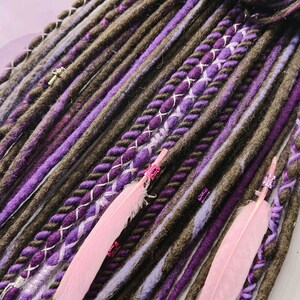 May include: A close-up of a set of brown and purple synthetic dreadlocks with pink feathers. The dreadlocks are braided with a variety of textures and colors, including brown, purple, and white.