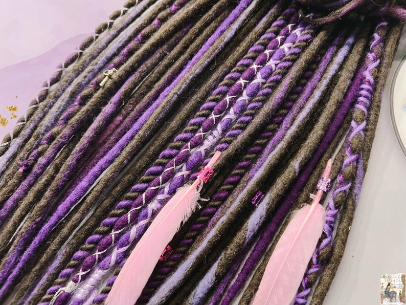 May include: A close-up of a set of brown and purple synthetic dreadlocks with pink feathers. The dreadlocks are braided with a variety of textures and colors, including brown, purple, and white.