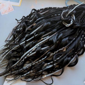 May include: Black and gray dreadlocks with wavy texture, adorned with small decorative accents. The dreadlocks are arranged in a layered style, with some sections braided. The overall look is dark and textured, suitable for alternative fashion.