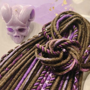 May include: A close-up of a bundle of brown and purple synthetic dreadlocks. The dreadlocks are made of a soft, textured yarn and have a subtle ombre effect.