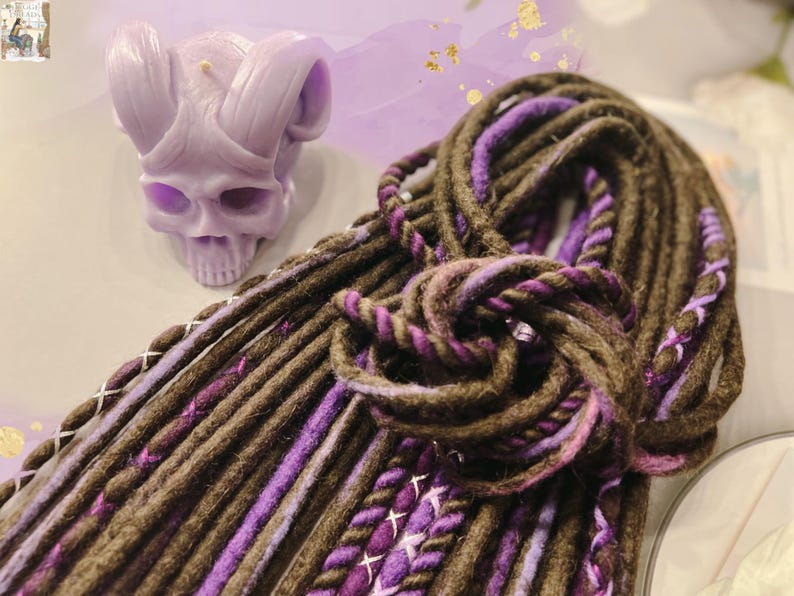 May include: A close-up of a bundle of brown and purple synthetic dreadlocks. The dreadlocks are made of a soft, textured yarn and have a subtle ombre effect.