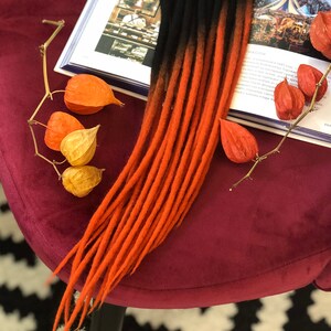 May include: A set of long, ombre hair extensions with black and orange strands. The extensions are lying on a burgundy velvet surface with dried orange and yellow flowers.