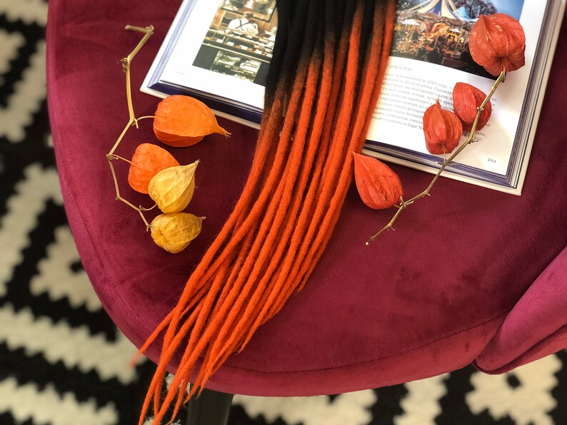 May include: A set of long, ombre hair extensions with black and orange strands. The extensions are lying on a burgundy velvet surface with dried orange and yellow flowers.