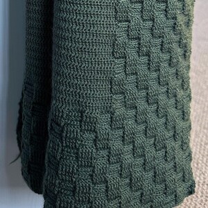 May include: A close-up of a dark green knitted blanket with a woven basket weave pattern. The blanket is folded over, revealing the texture and depth of the knit. The color is a deep, earthy green.